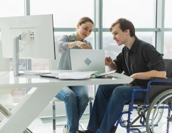 Dos personas trabajando juntas frente a un portátil en una oficina, una de ellas tiene una silla de ruedas